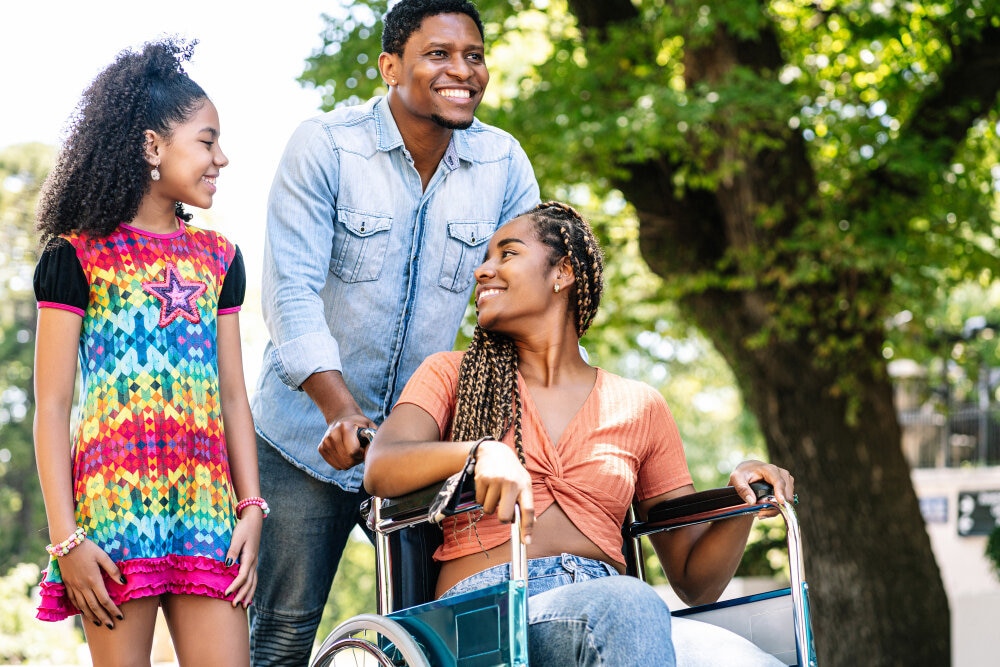 A man standing behind a smiling young woman who uses a wheelchair, outdoors among trees.