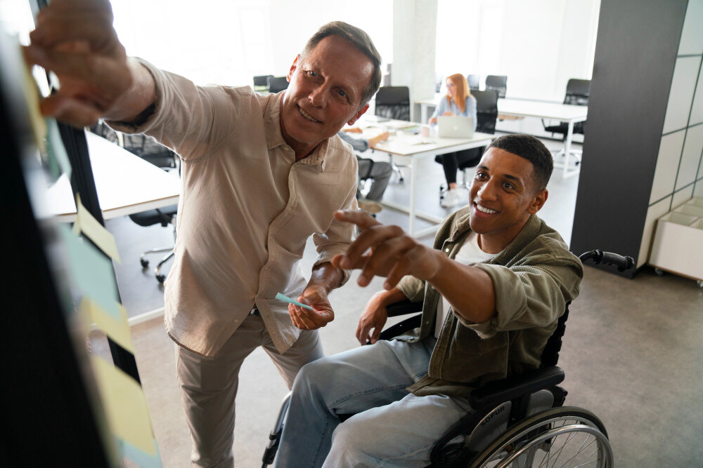 Two colleagues working through notes on a board, one of them using a wheelchair.