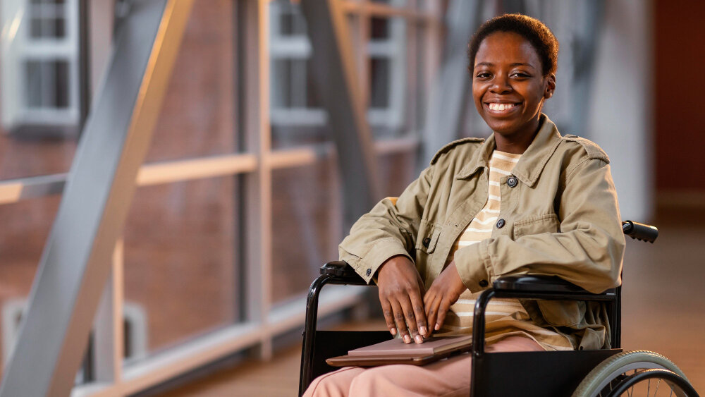 A smiling woman using a wheelchair, holding a folder in a bright building.