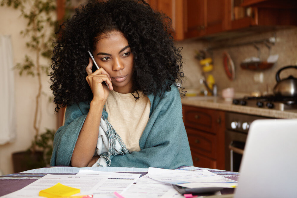 A woman at her kitchen table on the phone, paperwork spread out in front of her.